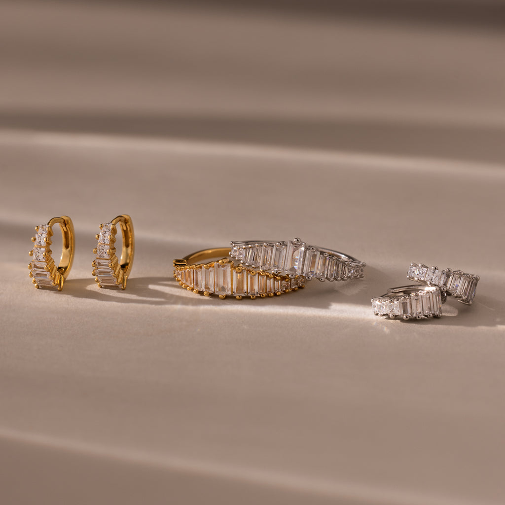 Gold and silver Art Deco Diamond Earrings & Ring Set with rectangular gemstones evoke vintage glamour, elegantly arranged on a soft beige surface with gentle shadows.