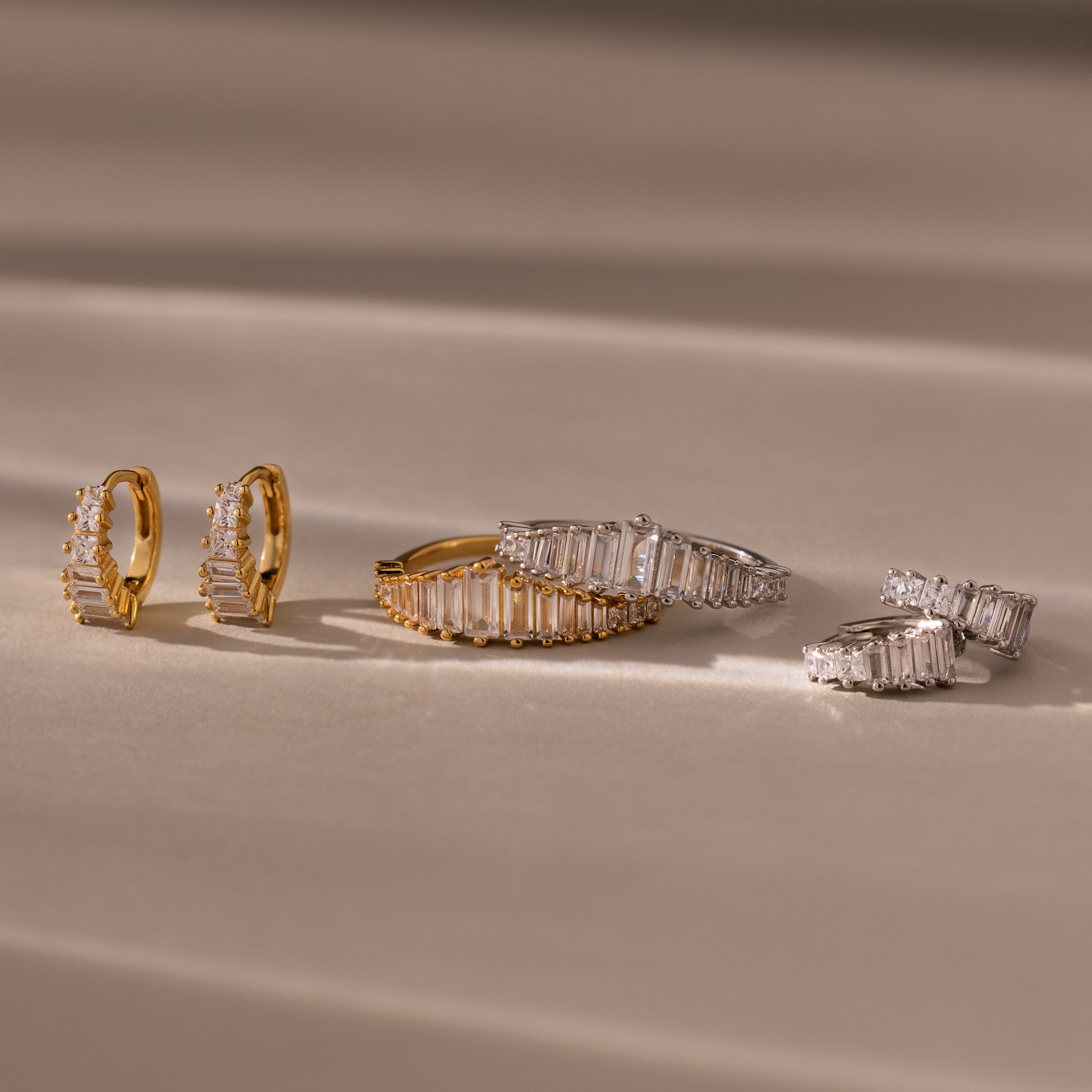 Gold and silver Art Deco Diamond Earrings & Ring Set with rectangular gemstones evoke vintage glamour, elegantly arranged on a soft beige surface with gentle shadows.