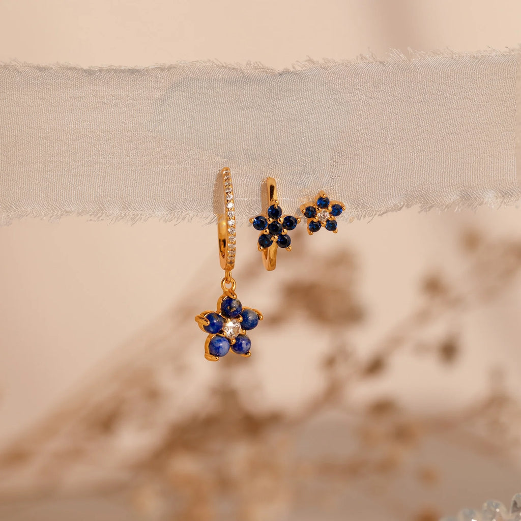 The Sapphire Flower Earrings Set features gold hoops with blue gemstone flower charms, elegantly displayed on sheer fabric against a soft, neutral background—a stunning jewelry addition.