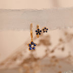 The Sapphire Flower Earrings Set features gold hoops with blue gemstone flower charms, elegantly displayed on sheer fabric against a soft, neutral background—a stunning jewelry addition.