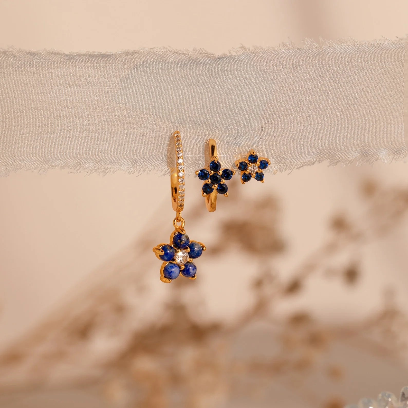The Sapphire Flower Earrings Set features gold hoops with blue gemstone flower charms, elegantly displayed on sheer fabric against a soft, neutral background—a stunning jewelry addition.