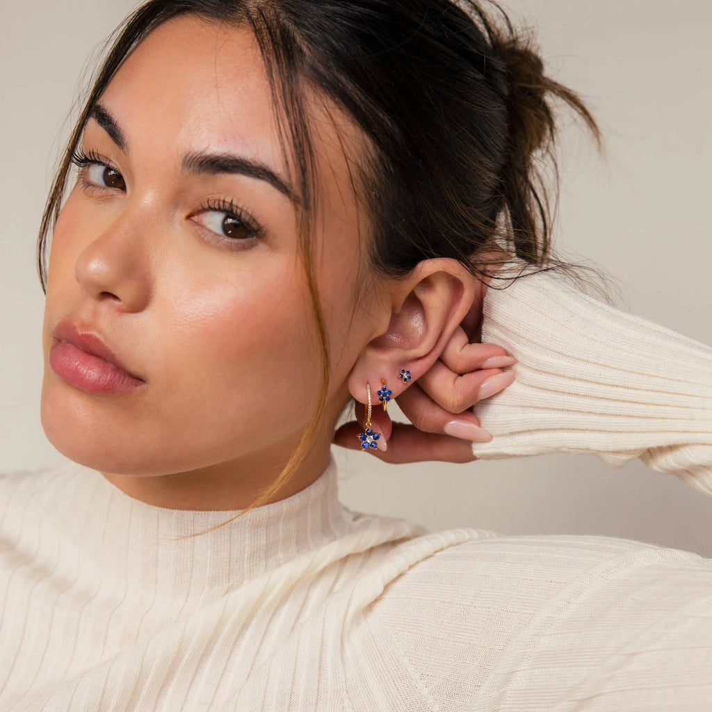 A woman in a cream ribbed turtleneck sweater touches her ear, showcasing the Sapphire Flower Earrings Set while looking at the camera.