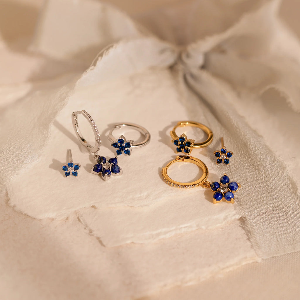 2 pairs of our Sapphire Flower Earrings Set in gold and silver on a beige fabric background