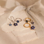 2 pairs of our Sapphire Flower Earrings Set in gold and silver on a beige fabric background