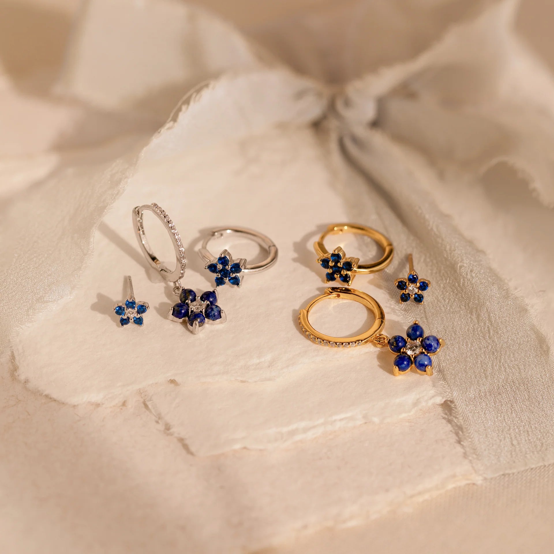 2 pairs of our Sapphire Flower Earrings Set in gold and silver on a beige fabric background