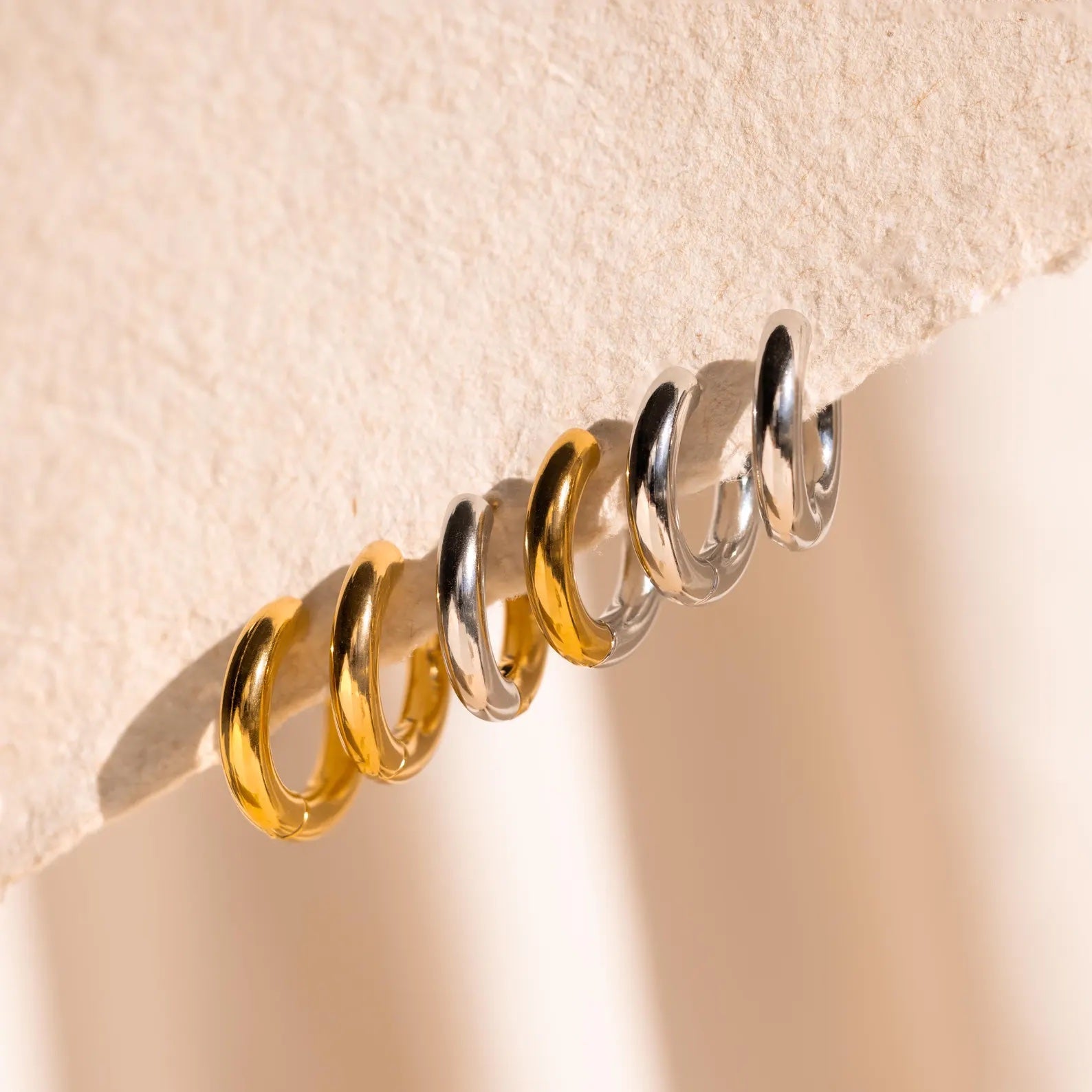 Three Mixed Metal Chunky Hoops in gold and silver are displayed on a textured beige surface, highlighting the stylish two-tone design of these hoop earrings.