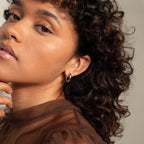 A woman with curly hair poses with her hand near her chin, wearing a sheer brown top and showcasing the Mixed Metal Chunky Hoops.