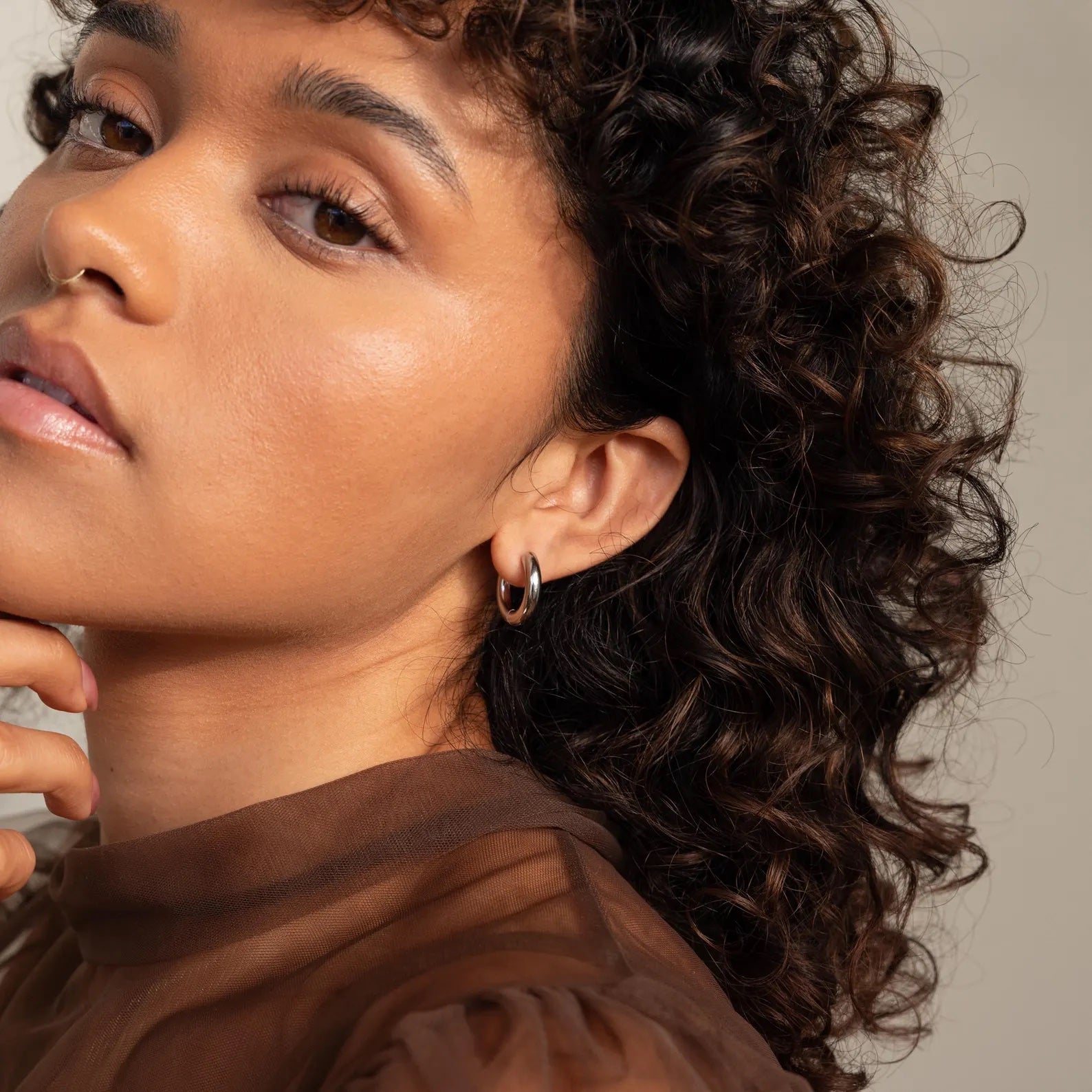 A woman with curly hair poses with her hand near her chin, wearing a sheer brown top and showcasing the Mixed Metal Chunky Hoops.