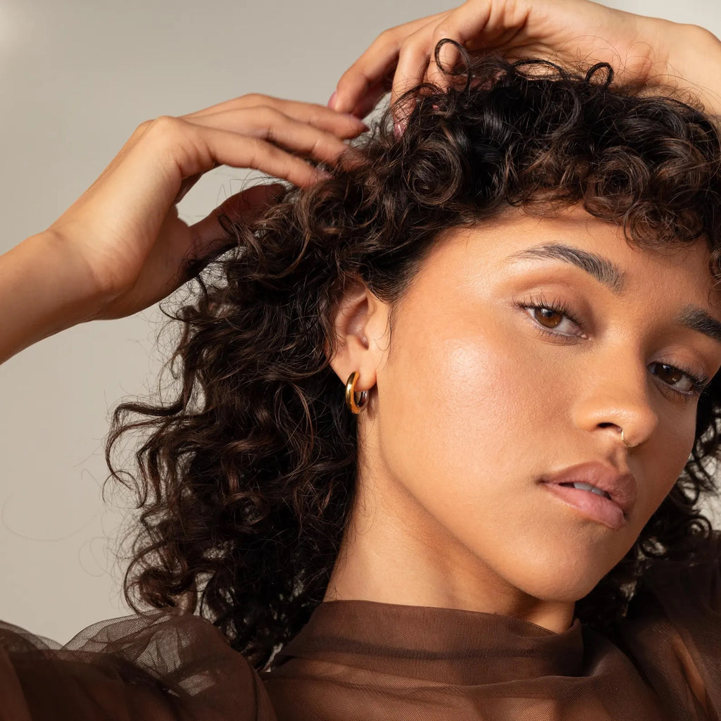 A woman with curly hair and gold hoop earrings poses in a sheer brown top, wearing stylish Mixed Metal Chunky Hoops and looking confidently at the camera.