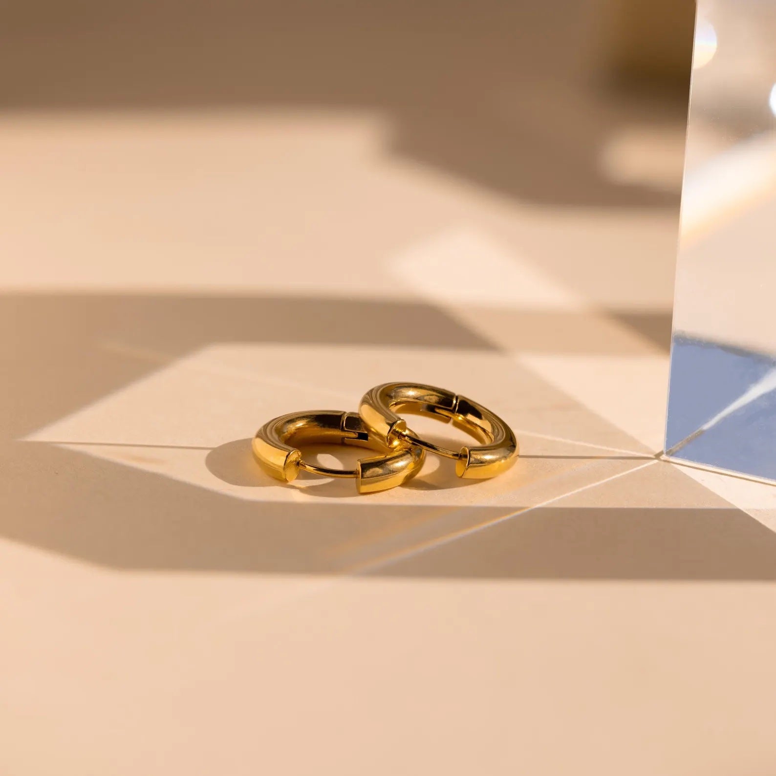 The Mixed Metal Chunky Hoops sit on a beige surface, where light and shadows cast striking geometric patterns around the bold earrings.