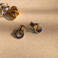 Mixed Metal Mini Triple Knot Earrings in gold and silver rest on a beige surface with soft shadows, capturing the effortless charm of everyday luxury.