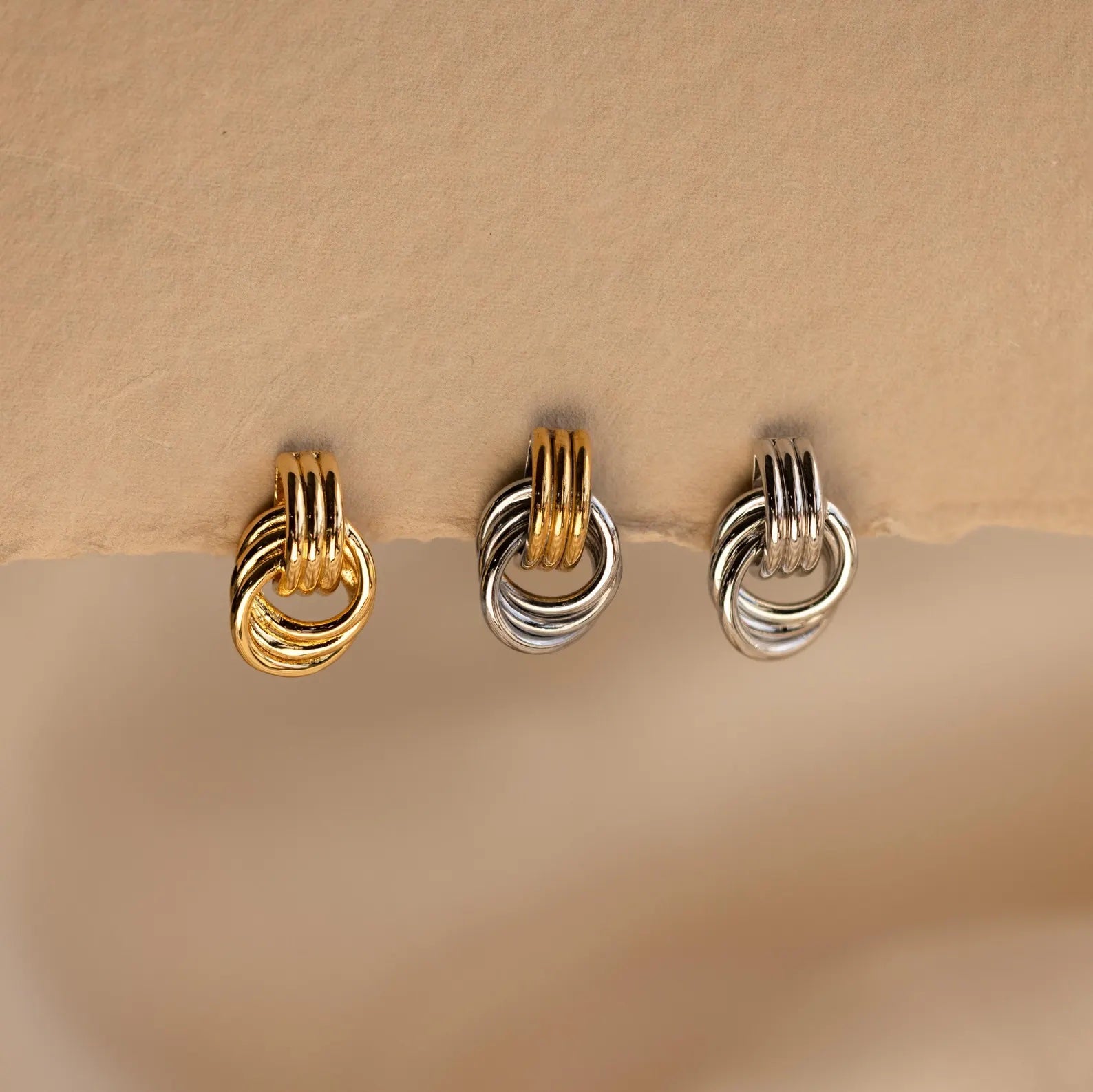 Three pairs of Mixed Metal Mini Triple Knot Earrings in gold, silver, and mixed tones rest on a beige surface—perfect for adding everyday luxury with these stunning earrings.