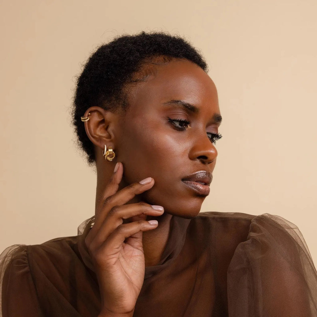 Woman with short hair wearing Mixed Metal Mini Triple Knot Earrings and a sheer brown top, posing with her hand near her face—embodying effortless everyday luxury.