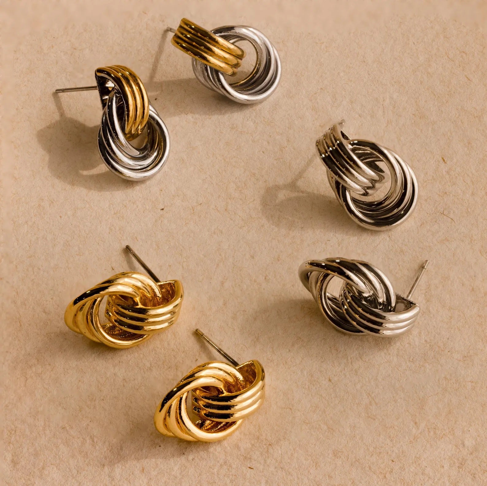 The Mixed Metal Mini Triple Knot Earrings—six gold and silver knot-shaped studs—are styled on a textured beige surface, offering a touch of everyday luxury to your collection.