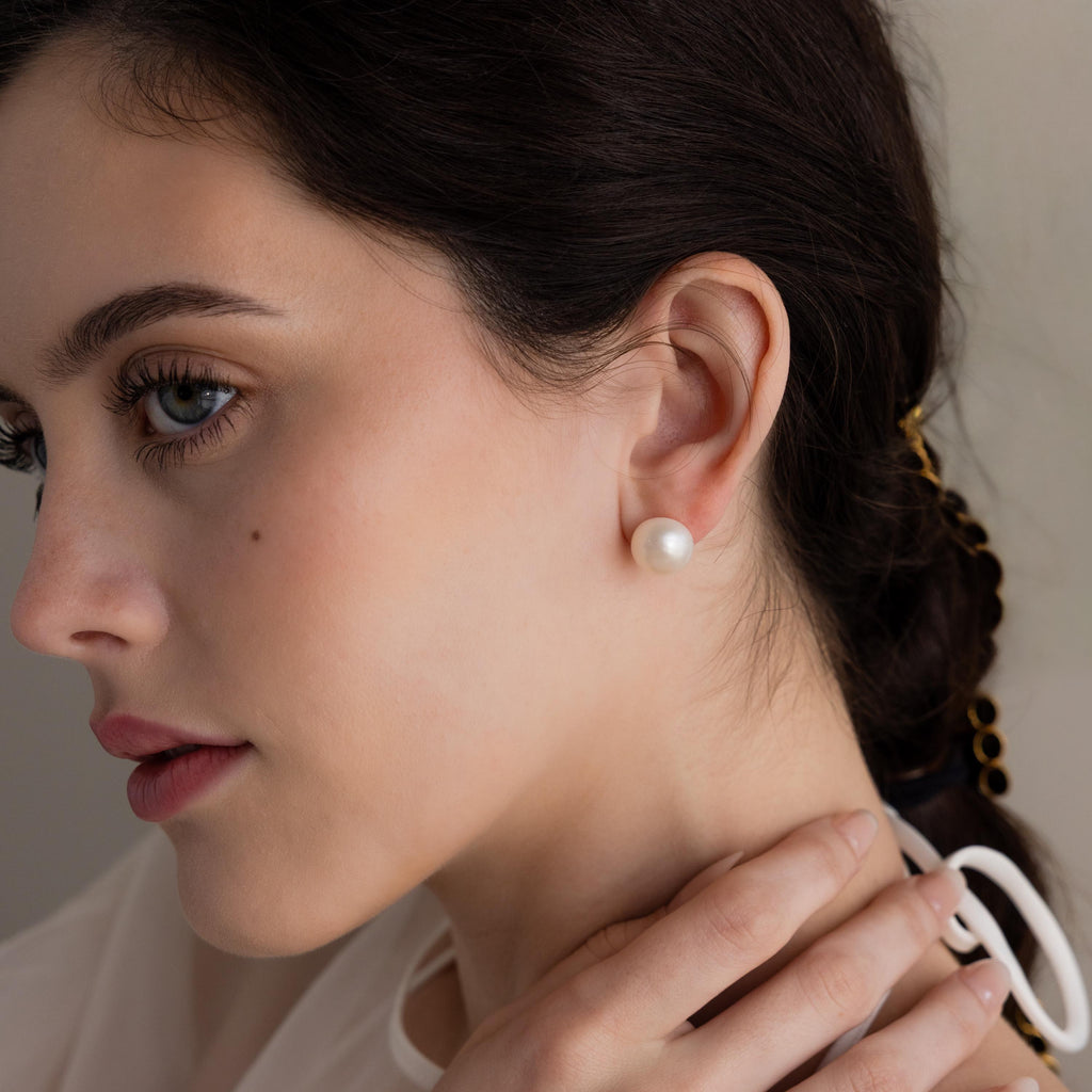Woman with dark hair wears Bold Pearl Studs—timeless white pearl earrings—gazing to the side with her hand near her neck.