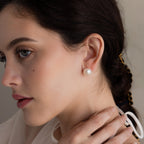 Woman with dark hair wears Bold Pearl Studs—timeless white pearl earrings—gazing to the side with her hand near her neck.
