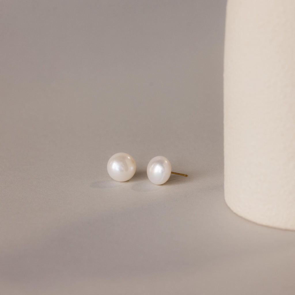 A pair of Bold Pearl Studs rests on a neutral surface next to a textured cream cylinder, highlighting the timeless elegance of this jewelry.
