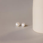 A pair of Bold Pearl Studs rests on a neutral surface next to a textured cream cylinder, highlighting the timeless elegance of this jewelry.