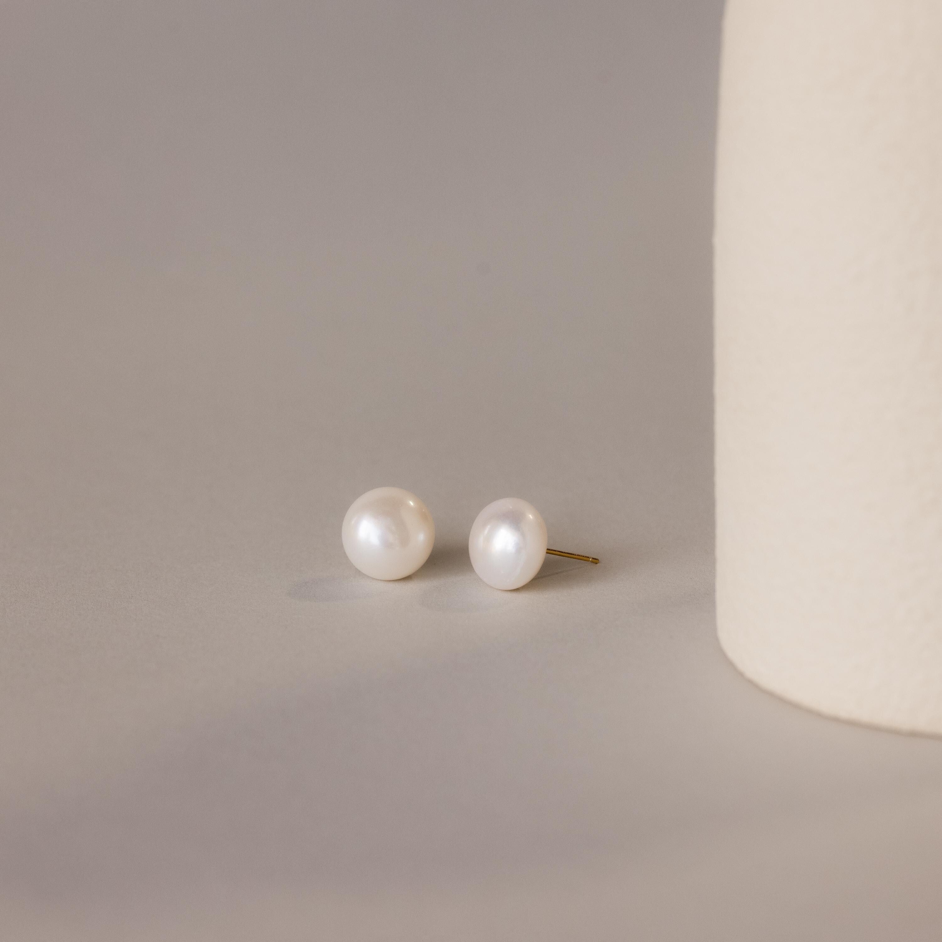A pair of Bold Pearl Studs rests on a neutral surface next to a textured cream cylinder, highlighting the timeless elegance of this jewelry.