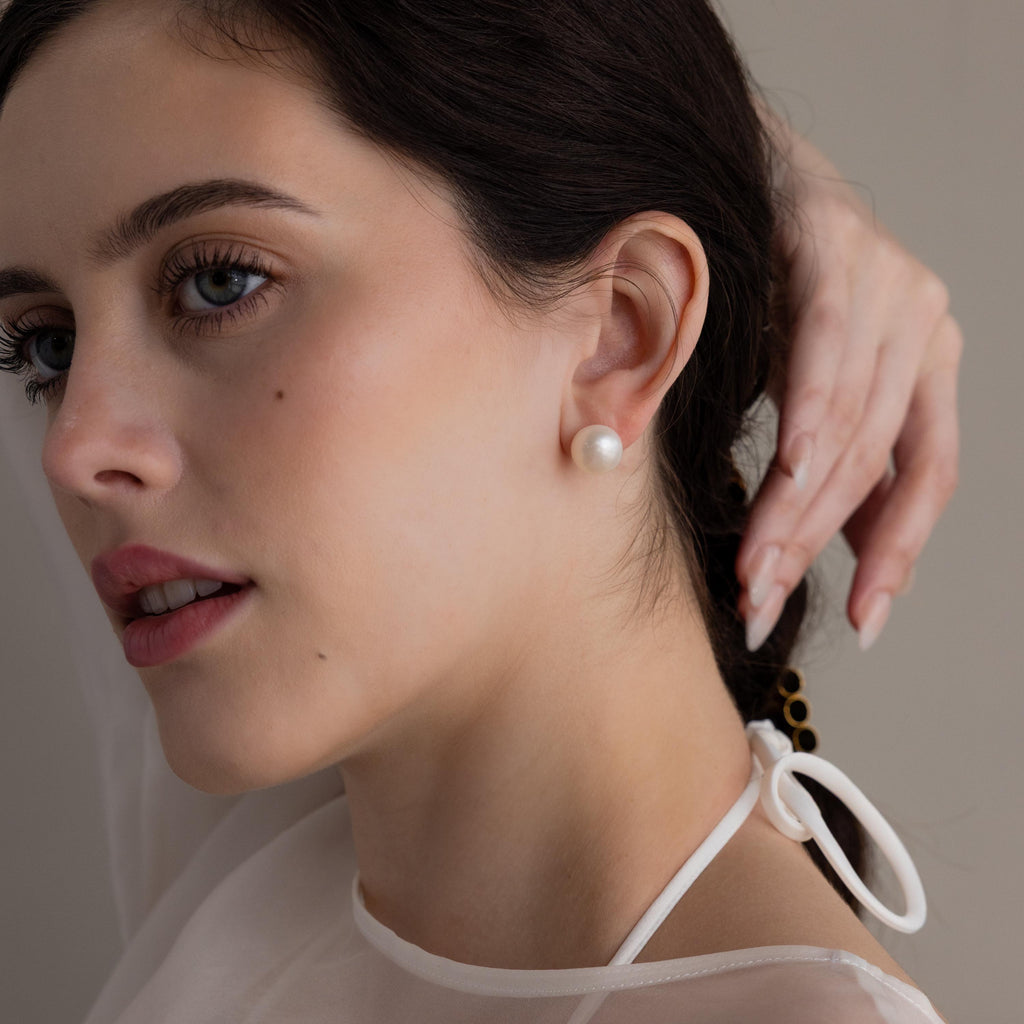 A fair-skinned woman wearing Bold Pearl Studs and a white top touches her dark hair while gazing to the side.