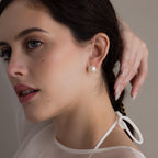 A fair-skinned woman wearing Bold Pearl Studs and a white top touches her dark hair while gazing to the side.