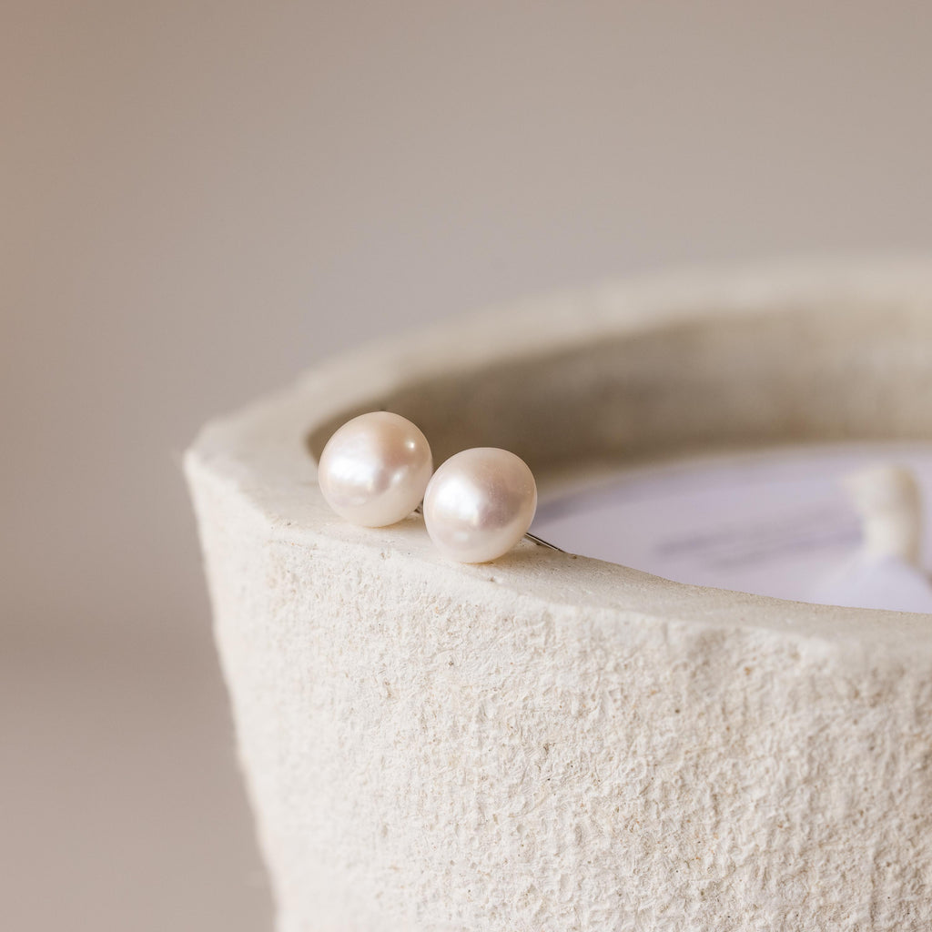 Two Bold Pearl Studs sit atop the textured rim of a beige cylindrical container, showcasing timeless jewelry that brings subtle elegance to any ensemble.