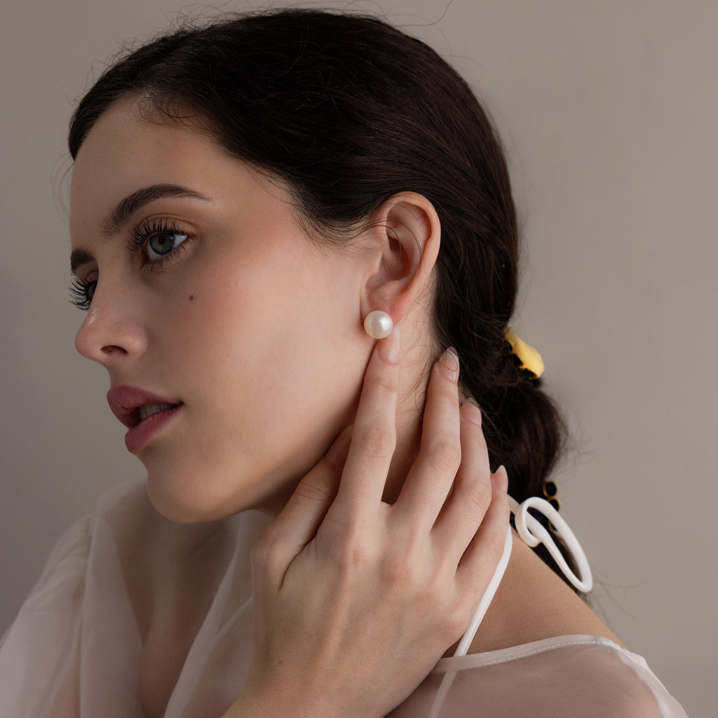 A woman with dark hair touches her Bold Pearl Studs while gazing to the side in a sheer white top—a graceful moment featuring classic, timeless jewelry.