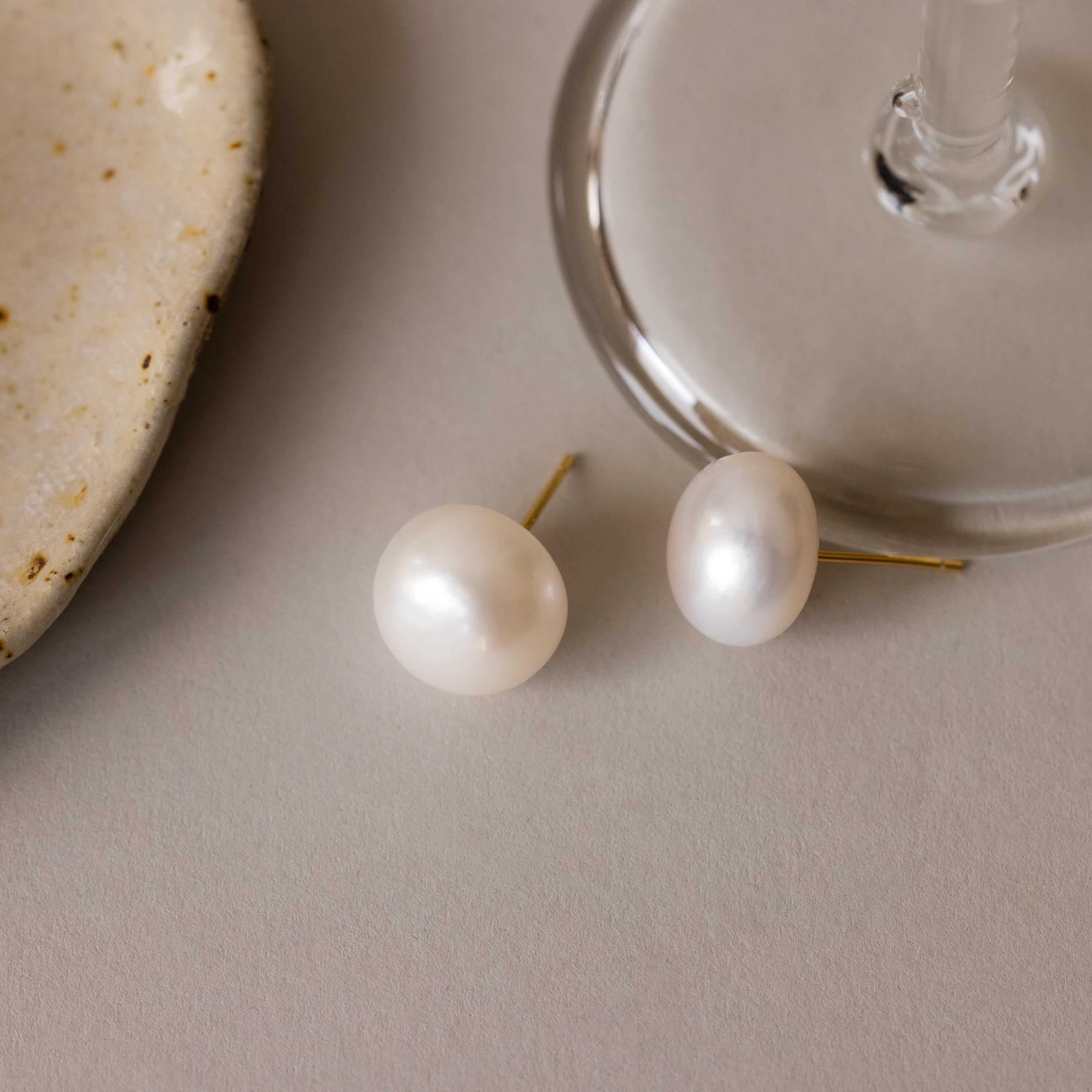 Bold Pearl Studs—elegant round pearl earrings with gold backs—rest on a neutral surface near a glass and stone, offering timeless jewelry style perfect for any occasion.