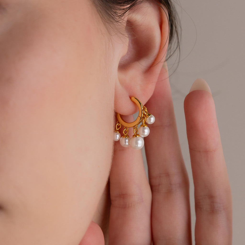 A hand touches an ear adorned with Pearl Chandelier Huggies, featuring gold hoops elegantly detailed with dangling pearl beads.