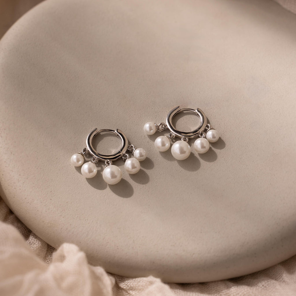Pearl Chandelier Huggies with delicate pearl beads dangle elegantly from silver hoops, displayed on a smooth beige surface.