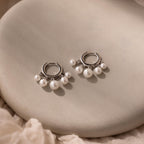 Pearl Chandelier Huggies with delicate pearl beads dangle elegantly from silver hoops, displayed on a smooth beige surface.