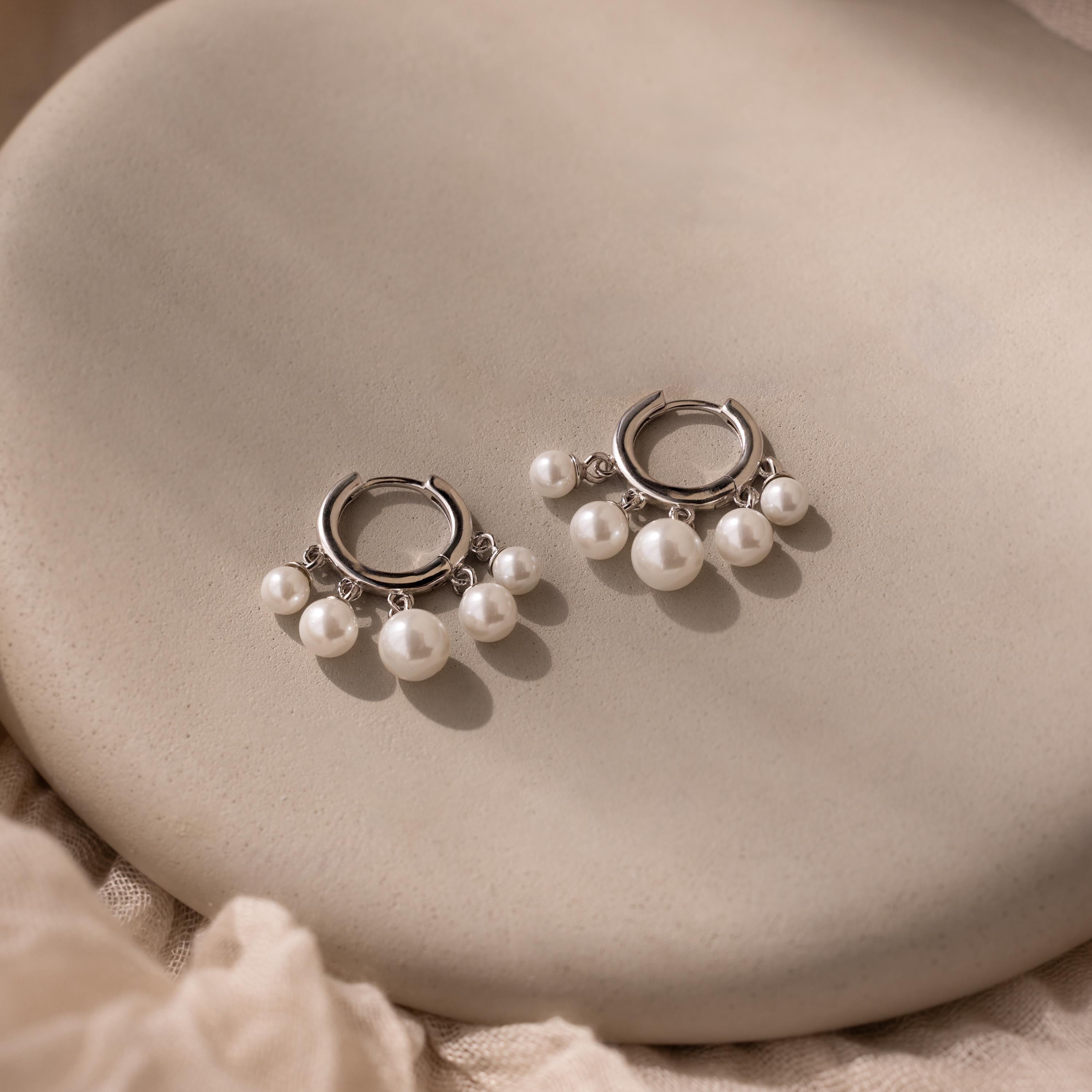 Pearl Chandelier Huggies with delicate pearl beads dangle elegantly from silver hoops, displayed on a smooth beige surface.