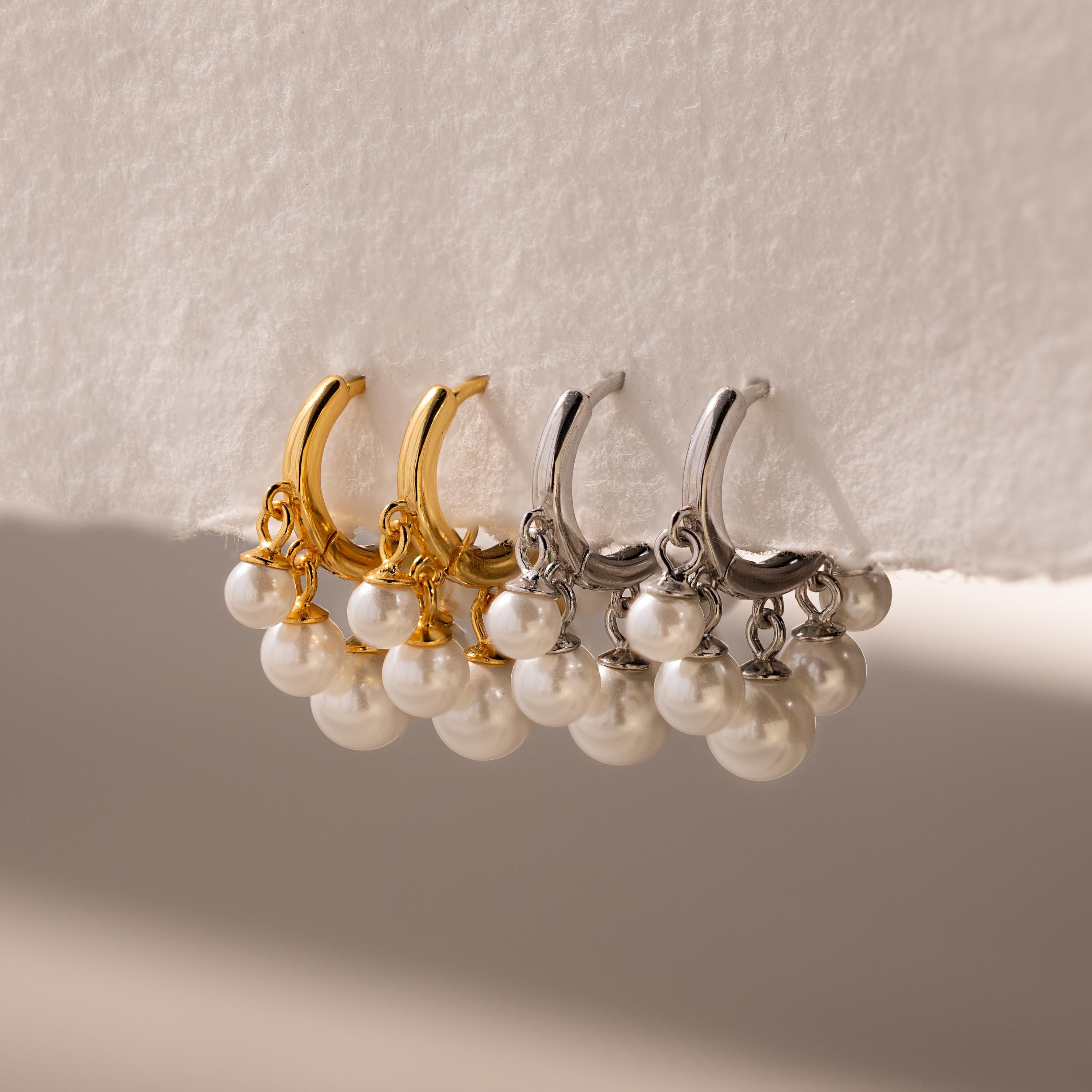 Two pairs of Pearl Chandelier Huggies, featuring gold and silver hoop earrings adorned with pearl beads, are elegantly displayed on a white surface.