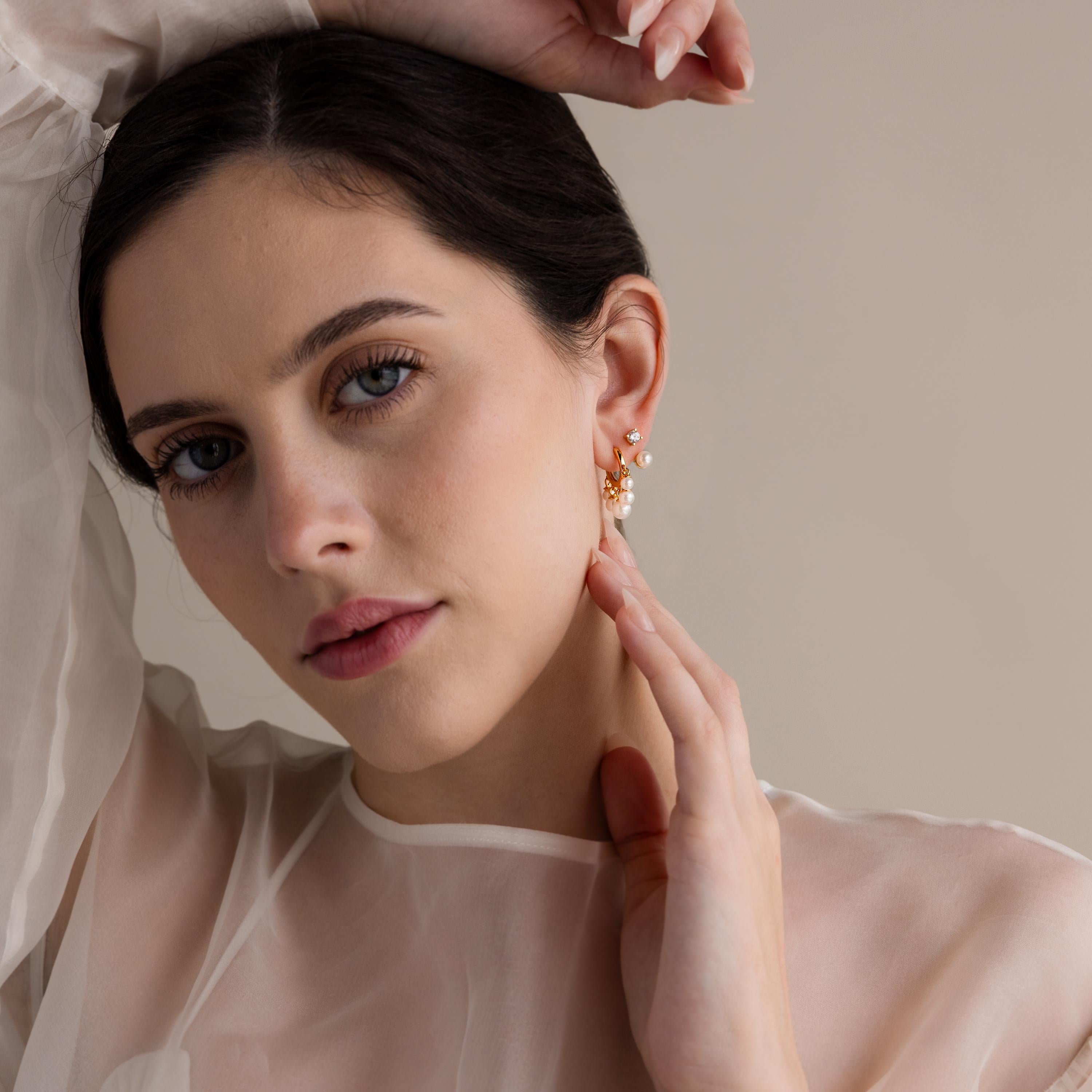 A woman in a sheer white top touches her ear, displaying elegant Pearl Chandelier Huggies—gold hoops adorned with dangling pearl beads—set against a neutral background.