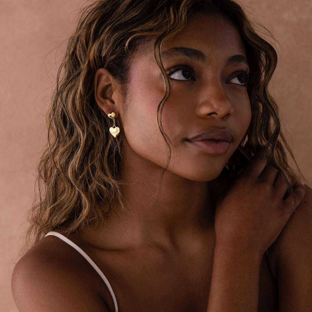 A woman with wavy hair wears Duo Heart Drop Studs and a white strap top, gazing thoughtfully to the side.