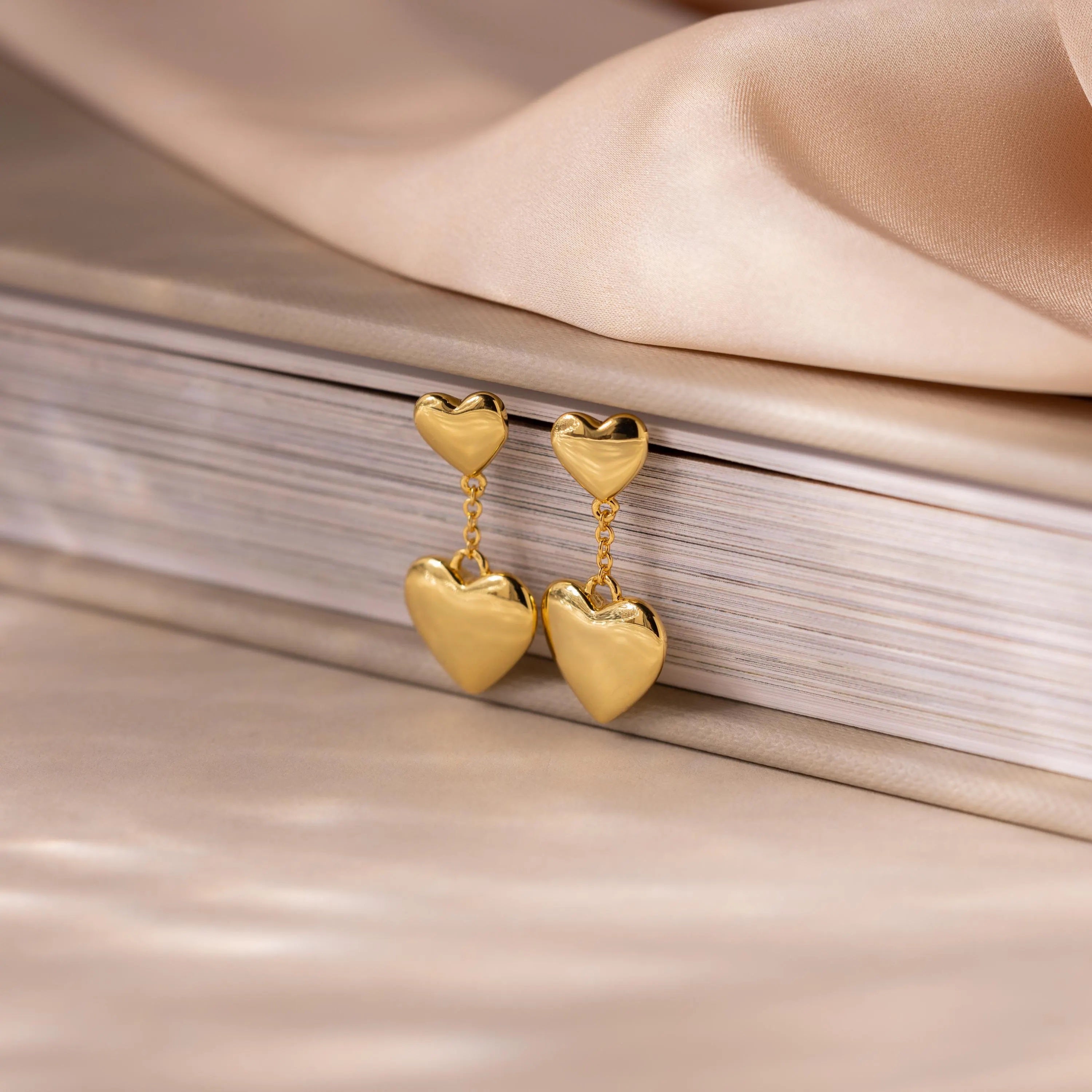 Duo Heart Drop Studs rest gracefully against a closed book on a soft beige fabric background.