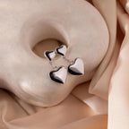 Duo Heart Drop Studs rest on a light stone, framed by beige satin—chic puffed heart earrings that add an elegant touch.