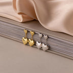 Duo Heart Drop Studs in gold and silver with a puffed heart design, shown against stacked books and beige fabric.