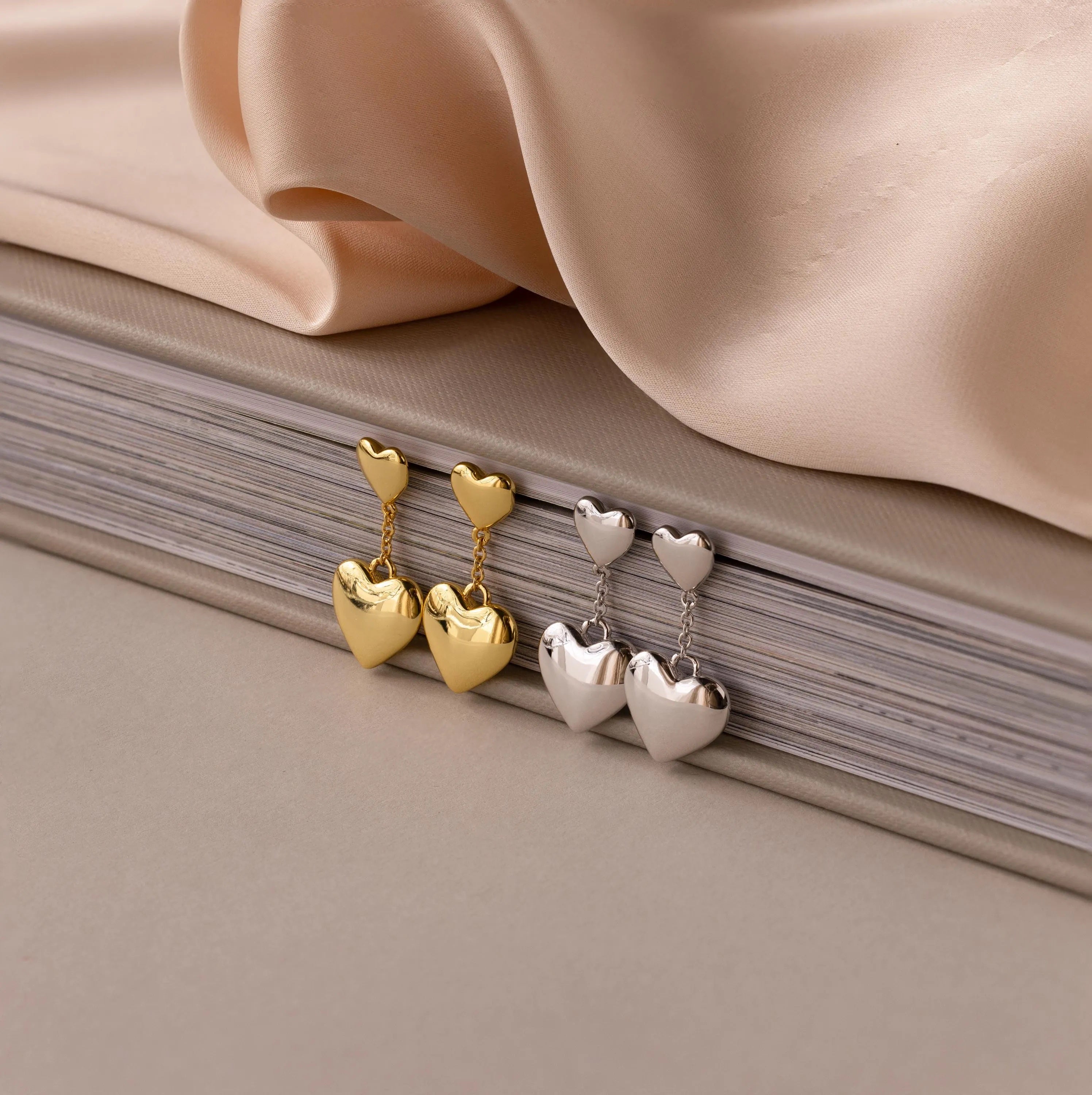 Duo Heart Drop Studs in gold and silver with a puffed heart design, shown against stacked books and beige fabric.