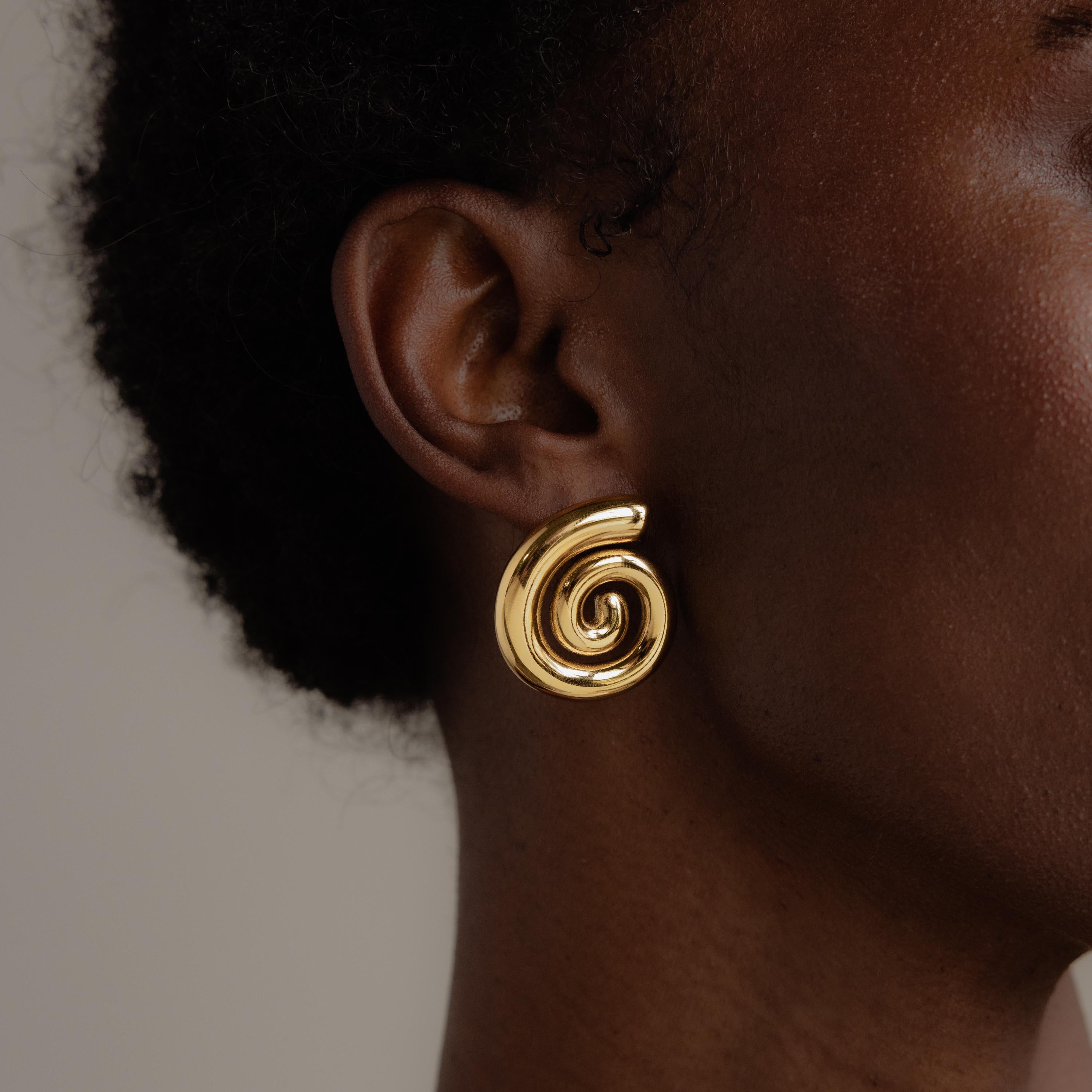 A close-up of a person wearing the Bold Spiral Stud Earrings—a striking gold geometric swirl—adorned on their ear.