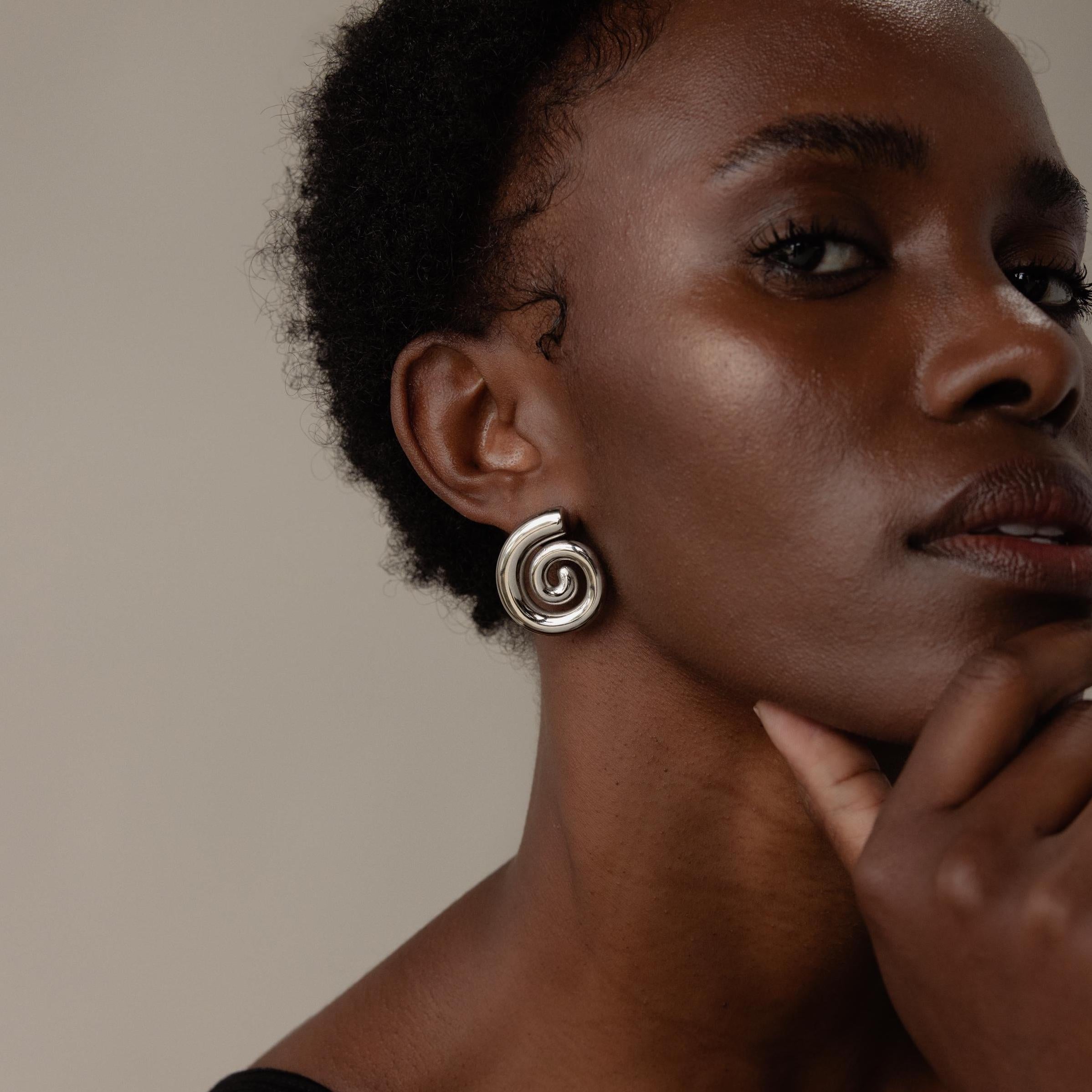 Woman with short hair wears Bold Spiral Stud Earrings, gazing thoughtfully with her hand near her chin.