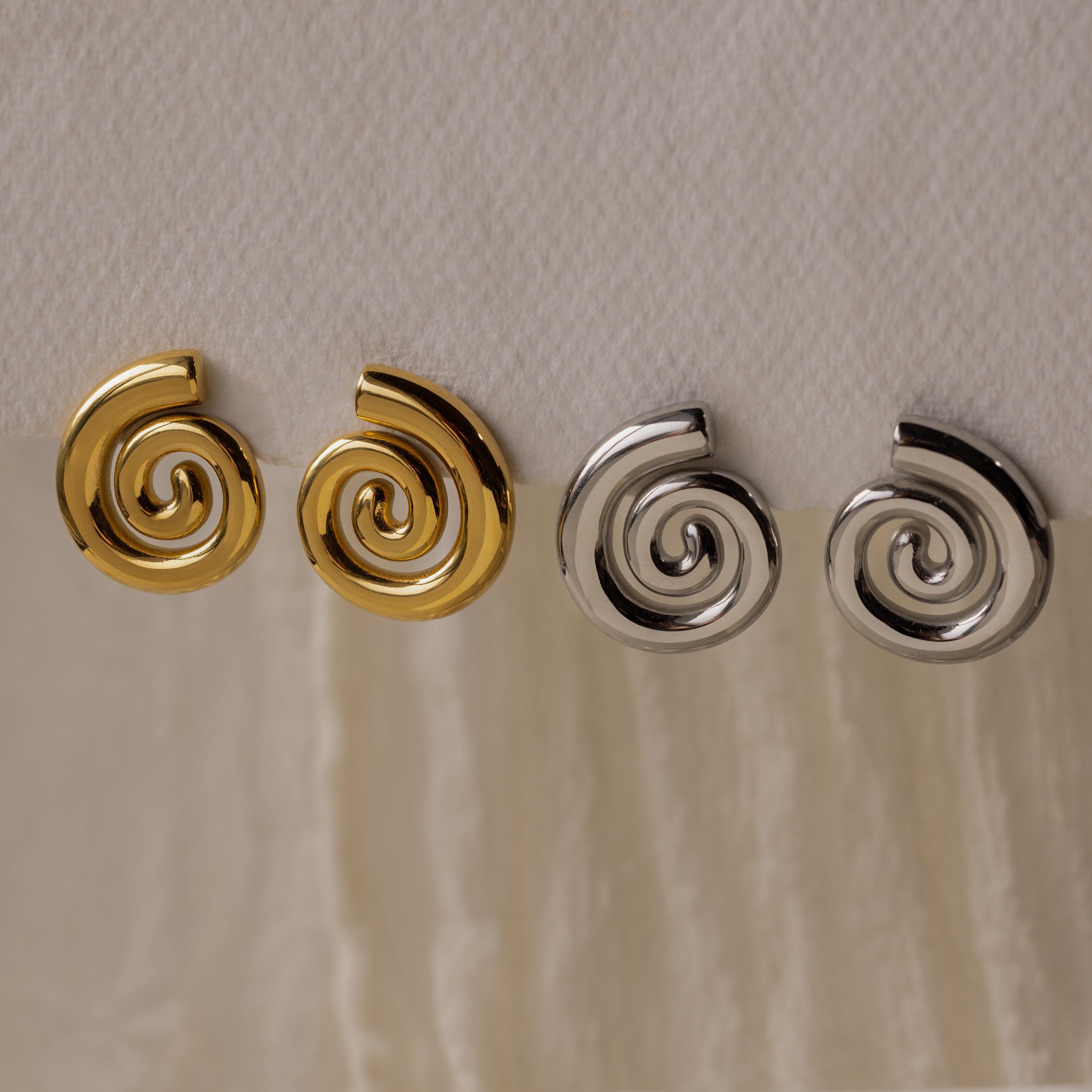 Two pairs of Bold Spiral Stud Earrings—one in gold and one in silver—are displayed on a white textured fabric, highlighting their unique geometric swirl design.