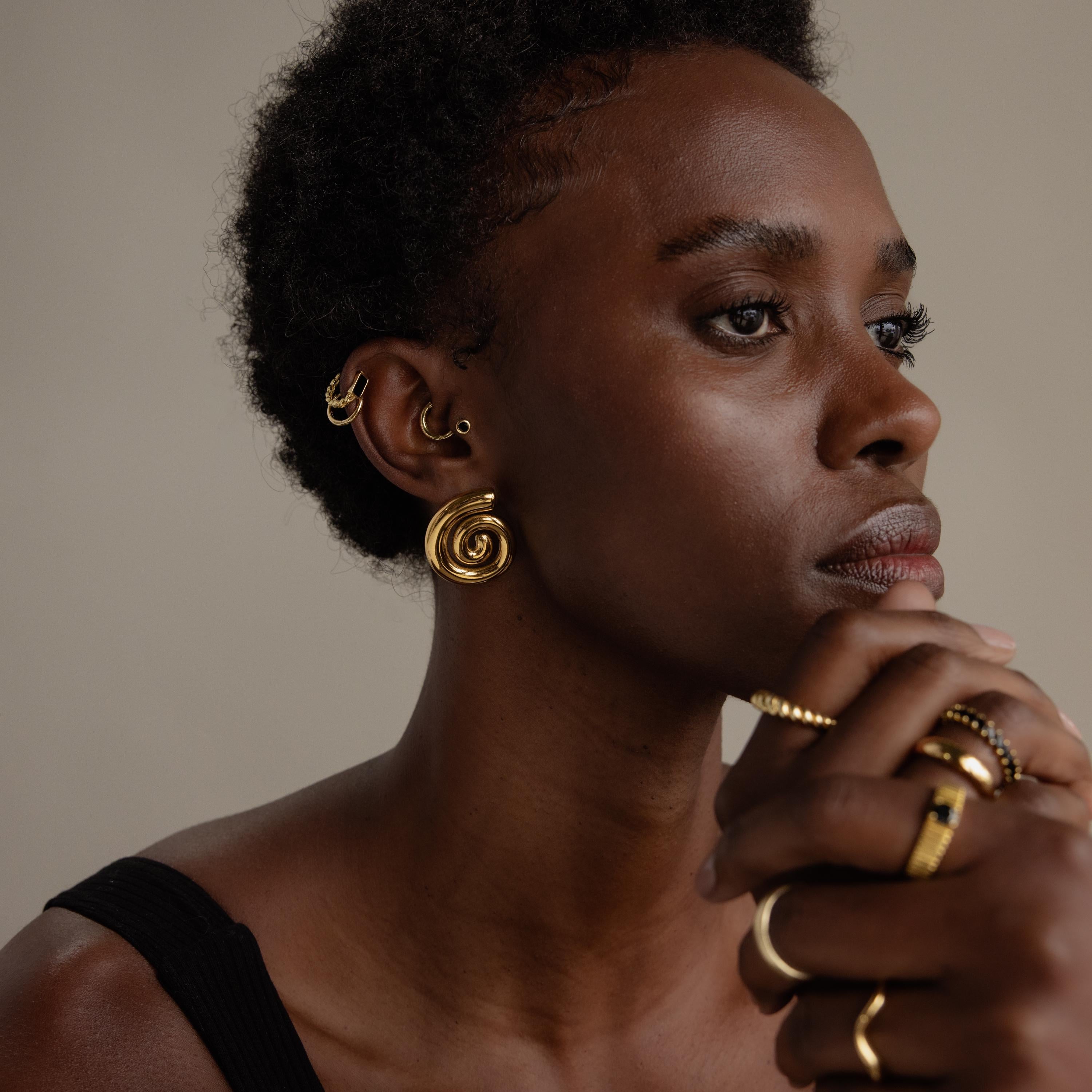 A woman wearing Bold Spiral Stud Earrings and multiple gold rings looks thoughtfully to the side.