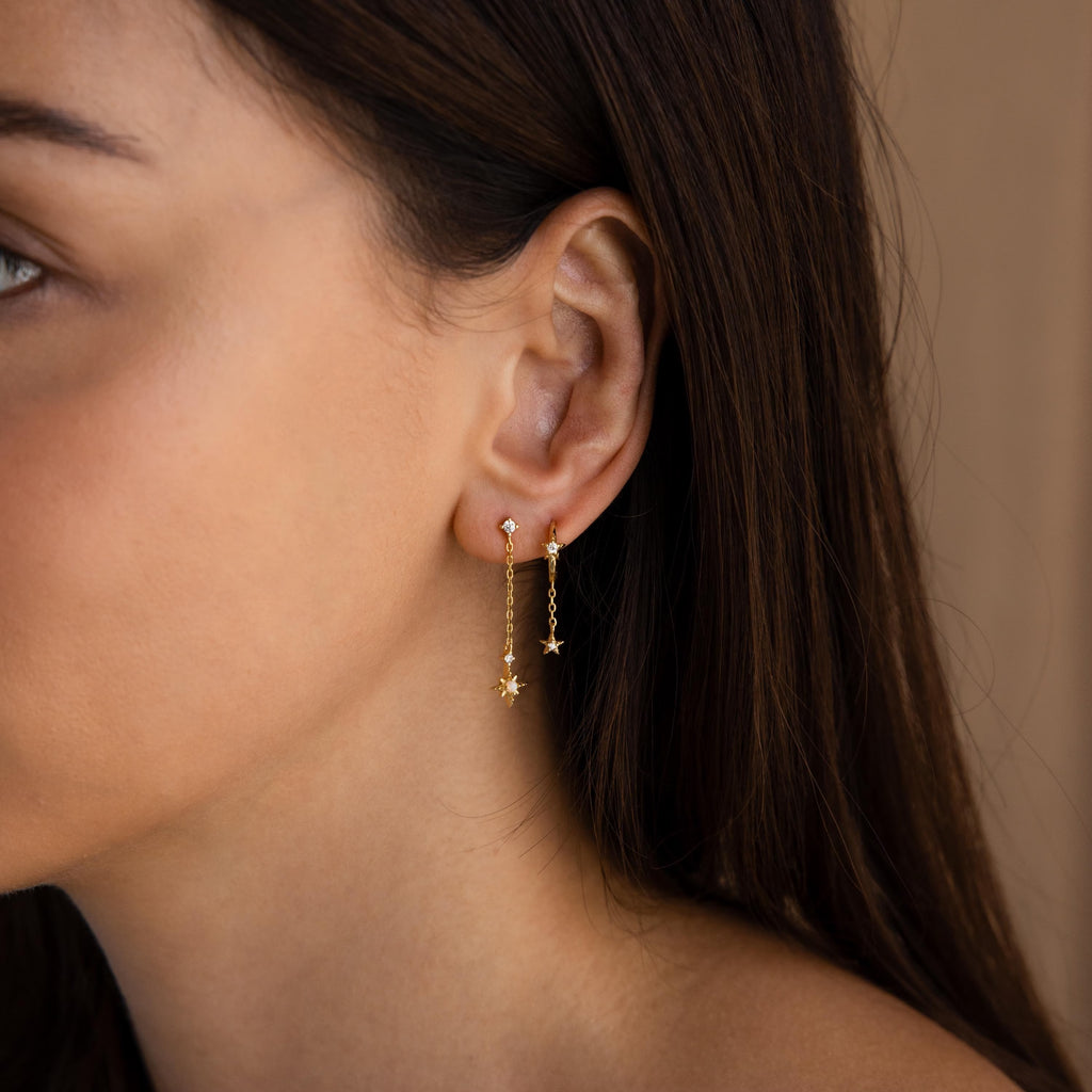 Model wears gold starburst chain stud earrings, accompanied by another star earring on a second piercing.