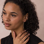 A woman with curly hair wears Agate Threader Earrings and a black top, gently touching her neck with a thoughtful expression.