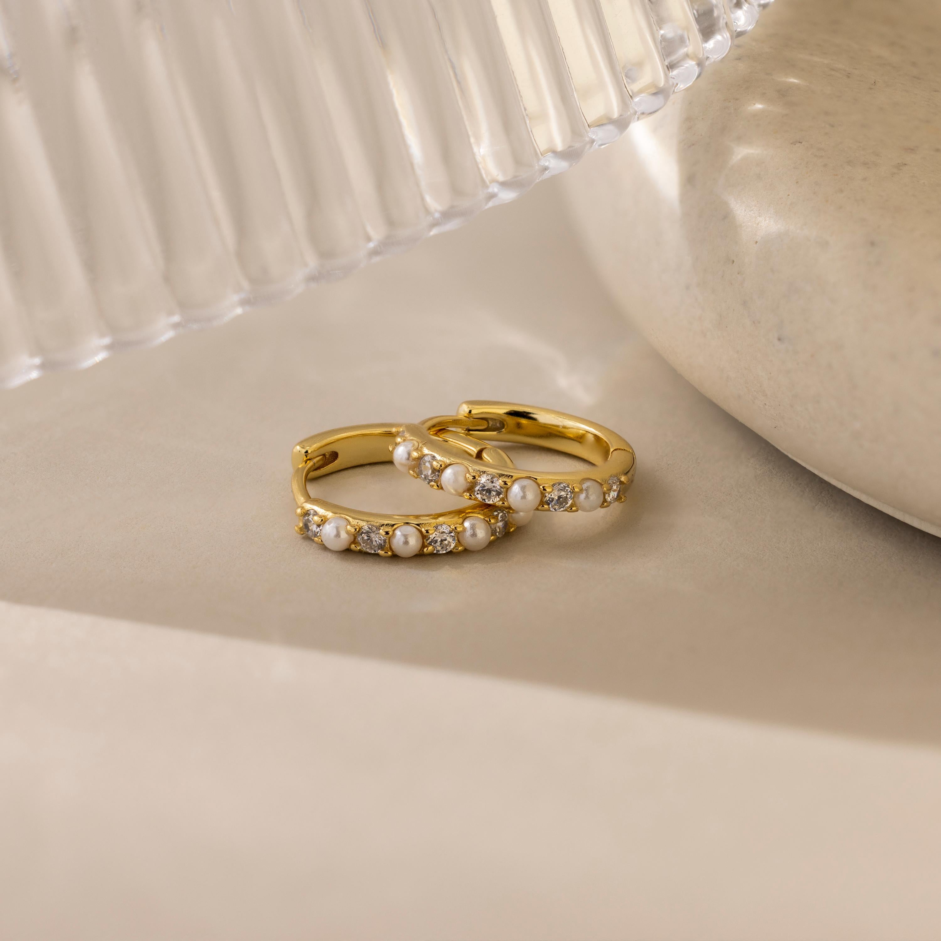 Pave Pearl Huggies are displayed on a beige surface, accented by nearby glass and ceramic objects.