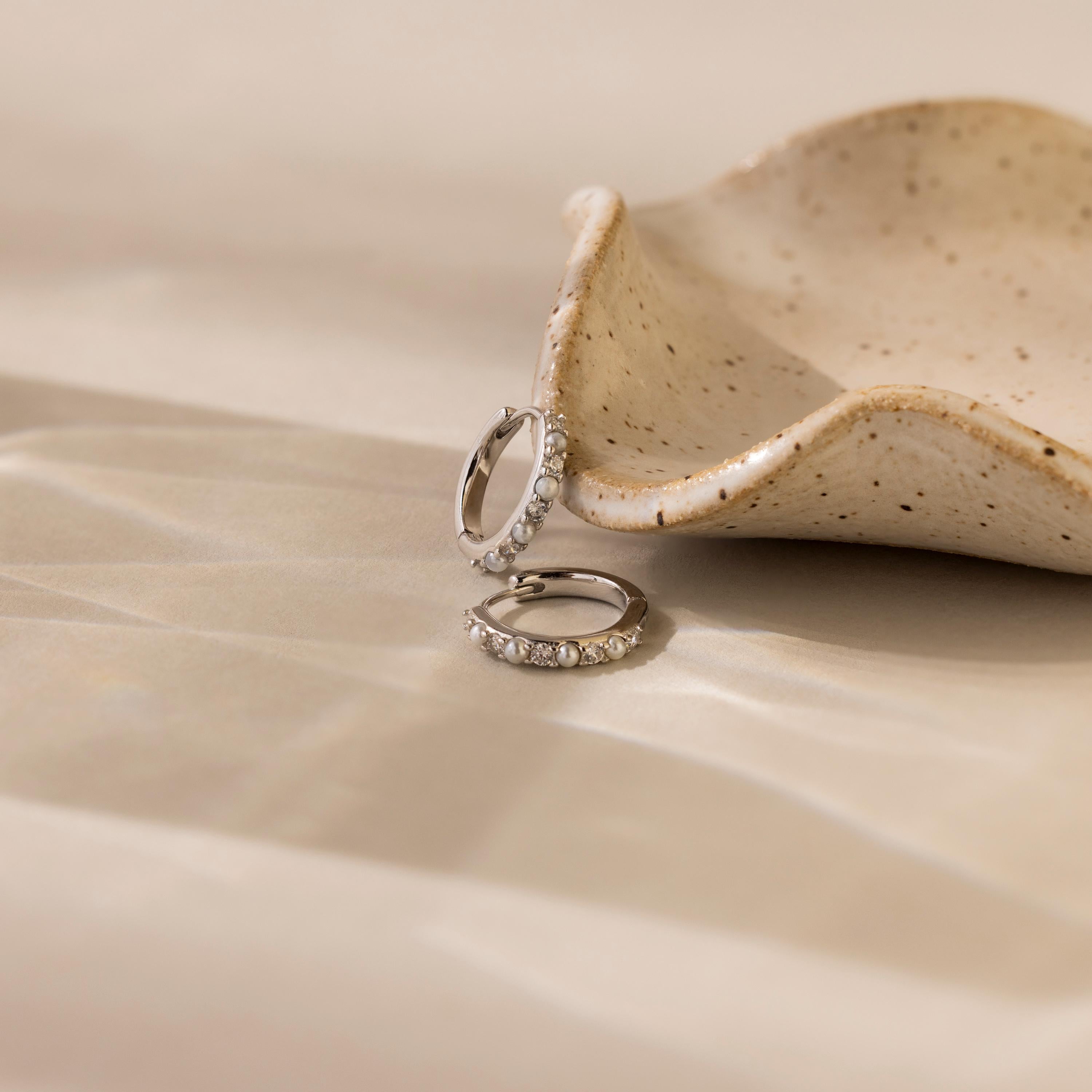 Two Pave Pearl Huggies rest beside a speckled ceramic dish on a beige surface.