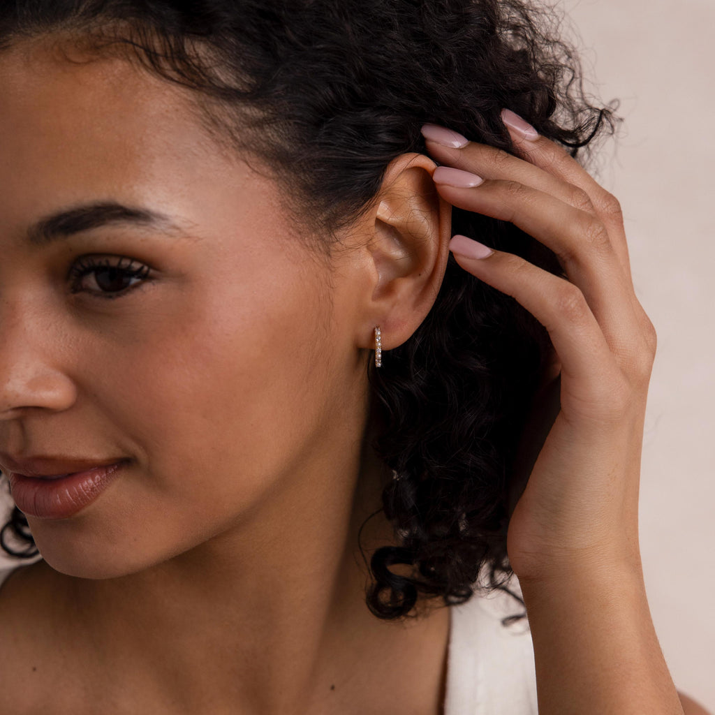 Woman with curly hair touches her ear, displaying Pave Pearl Huggies and neutral nail polish.