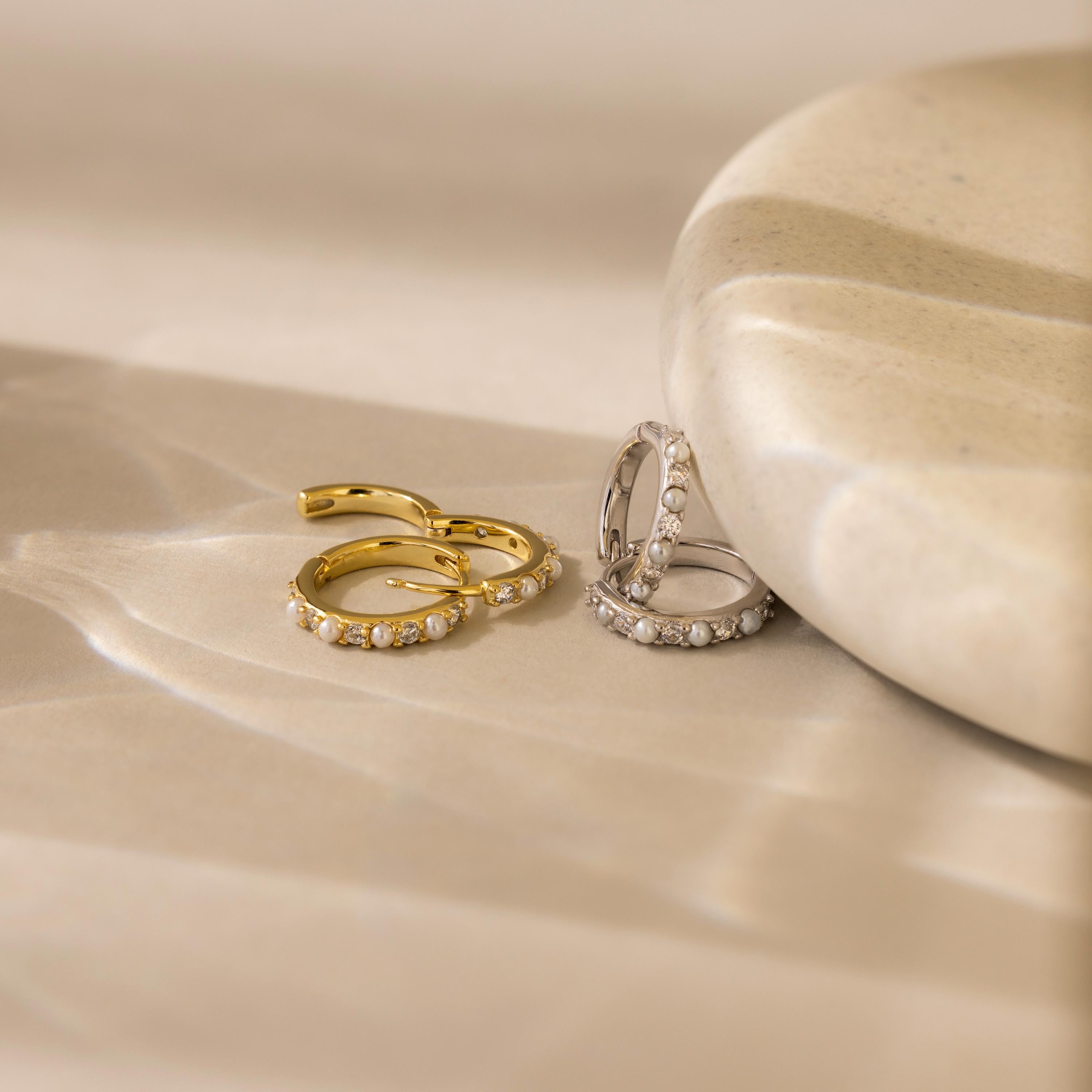 The Pave Pearl Huggies shine on a beige surface with soft shadows, joined by gold and silver diamond-studded rings and elegant accents for a refined look.