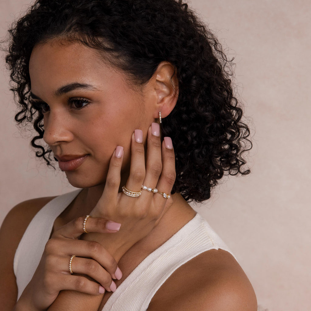 A woman with curly hair wears gold rings and Pave Pearl Huggies earrings, posing with her hands near her face against a neutral background.